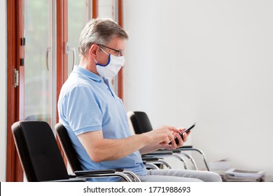 Mature Man With Face Mask Sitting In A Bright Waiting Room Looking At Smartphone