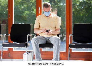 Mature Man With Face Mask Sitting In A Bright Waiting Room Of A Hospital Or An Office Looking At Smart Phone