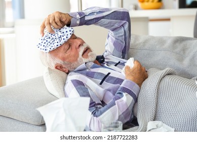 Mature Man Feeling Sick With Cold And Fever At Home, Ill With Flu Disease Sitting On The Sofa With Ice Pack On His Head