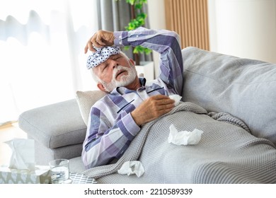 Mature Man Feeling Sick With Cold And Fever At Home, Ill With Flu Disease Sitting On The Sofa With Ice Pack On His Head