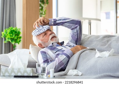 Mature Man Feeling Sick With Cold And Fever At Home, Ill With Flu Disease Sitting On The Sofa With Ice Pack On His Head