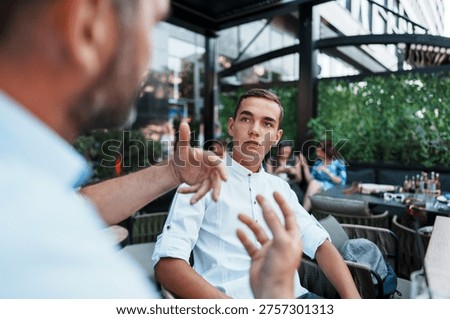 A mature man gestures while talking to his teenage son who listens attentively during a serious discussion at an outdoor restaurant terrace.