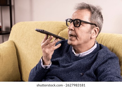 Mature man in glasses dictating a voice message to his smartphone voice assistant while relaxing on a sofa at home. Concept of accessibility, hands-free communication, modern technology for seniors. - Powered by Shutterstock - Get 15% off with code: PIKWIZARD15