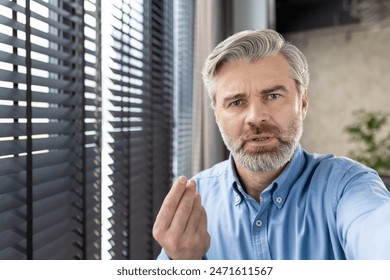 Mature man with gray hair and beard looking serious and gesturing near a window with blinds. Interior setting conveys a professional or personal communication. - Powered by Shutterstock