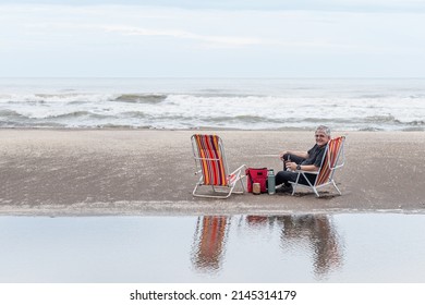 Mature Man With Gray Hair Sitting On A Beach Chair Smiling While Looking At The Camera. The Sea In The Background.