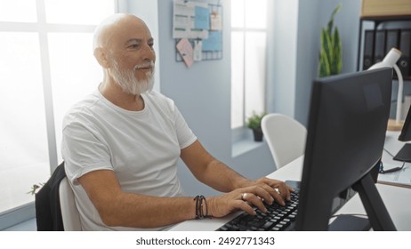 Mature man with grey hair and beard working on a computer in a bright office room with natural light, plants, and a bulletin board in the background - Powered by Shutterstock