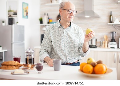 Mature Man Holding Green Apple Before Taking A Bite During Breakfast In Kitchen. Elderly Retired Person Eating Apple During Breakfast In Modern Cozy Kitchen. Healthy Green Happy Fresh Lifestyle