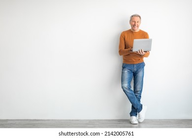 Mature Man Holding Using Laptop Standing Leaning On White Wall, Posing Looking At Camera. Happy Adult Working On Pc, Typing On Keyboard Isolated On Studio Background, Full Body Length, Free Copy Space