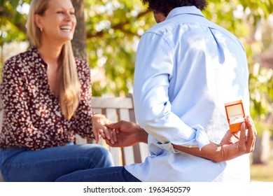 Mature Man Kneeling And Holding Engagement Ring Behind Back About to Propose To Woman In Park