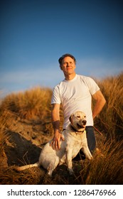 Mature Man Kneeling On A Grassy Sand Dune With His Dog.