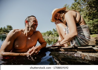 Mature Man In Lake With Woman Sitting At The Pier. Couple Enjoying On Their Camping Vacation At The Lake.