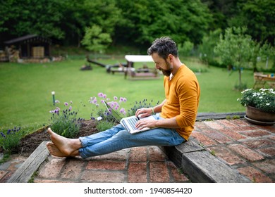 Mature Man With Laptop Working Outdoors In Garden, Green Home Office Concept.