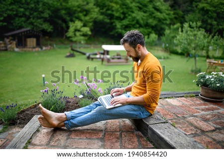 Mature man with laptop working outdoors in garden, green home office concept.