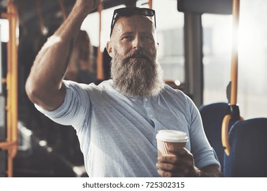 Mature man with a long beard smiling and drinking a takeaway cup of coffee while standing on a bus during his morning commute - Shutterstock ID 725302315