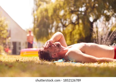 Mature Man Lying On Grass And Sunbathing At Home As Children Play In Pool