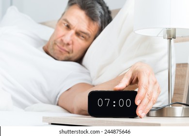 Mature Man On Bed Turning Off Clock On Nightstand