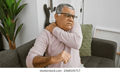 A mature man in a pink sweater sits indoors grimacing in pain, holding his shoulder with a guitar in the background. - Powered by Shutterstock