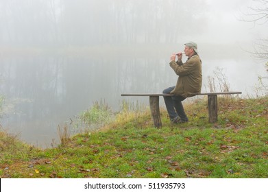 Mature man playing sopilka while sitting on a bench on the riverside at autumnal foggy morning - Powered by Shutterstock