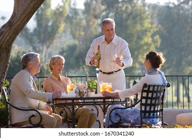 Mature Man Pouring Wine For Friends Dining At Outdoor Restaurant Table