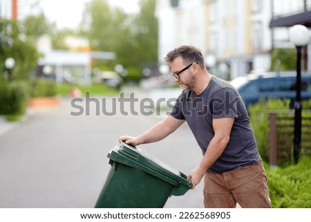 Mature man pulling out a large green plastic garbage container in front of the townhouse to the roadway of the street. Garbage collection and recycling trash, the zero waste concept. Waste managment
