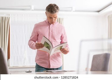 Mature Man Reading Brochure While Standing At Table In Apartment