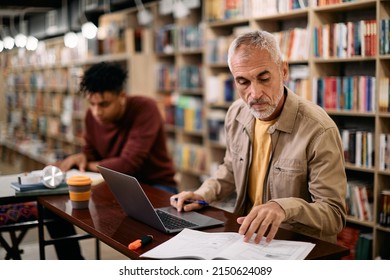 Mature man reading lecture notes and using laptop while studying in library. - Powered by Shutterstock