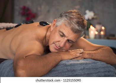 Mature man relaxing on bed during hot stone massage. Portrait of senior man with closed eyes resting during lastone therapy. Handsome man with black lava stone on back at spa with candles.