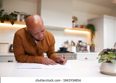 Mature Man Reviewing And Signing Domestic Finances And Investment Paperwork In Kitchen At Home