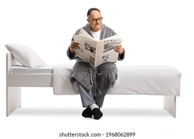 Mature Man In A Robe Relaxing On A Bed With A Newspaper Isolated On White Background