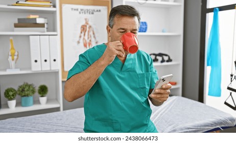 Mature man in scrubs drinking coffee and checking smartphone in a clinic's staff room. - Powered by Shutterstock