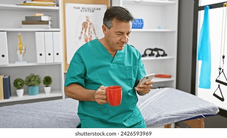 Mature man in scrubs sipping coffee and checking smartphone in a physiotherapy clinic interior. - Powered by Shutterstock