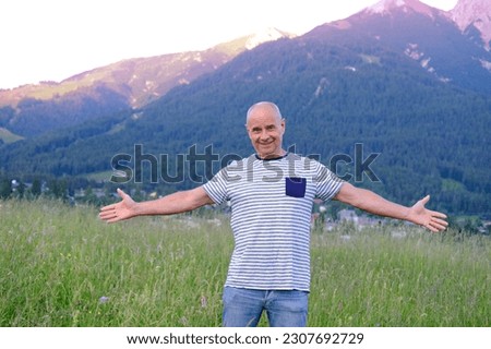 mature man, senior 60 years old standing in mountains above city, smiles, Alps are behind him, green grass in meadow, concept of enjoy life in old age, travel, active lifestyle, human happiness