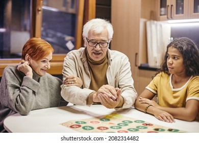 Mature man shaking dice in hands over board game while playing it with his wife and their cute interracial granddaughter