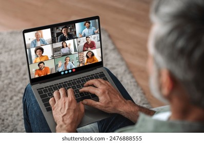 Mature man sits on a couch at home, using a laptop to participate in a virtual meeting. He is focused on the screen, which displays a grid of video feeds of other participants - Powered by Shutterstock