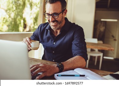 Mature Man Sitting At A Cafe Table Drinking Coffee And Using Laptop Computer. Man Doing His Work Sitting At A Coffee Shop.