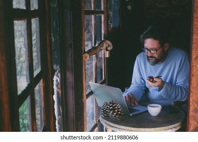 Mature man sitting at home working on laptop computer and smoking pipe. Alternative single lifestyle male people indoor in cozy cabin wooden chalet. Telecommuting online - Powered by Shutterstock