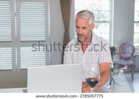 Mature man sitting at light table in bright room using laptop and sipping red wine. Homeoffice, naturallight, minimalist, interior, windows, armchair, beverage