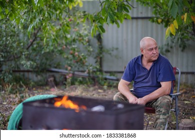 Mature Man Sitting On Chair In Garden. Older Male Lit Fire In Chargrill For Roasting Shashlik With Vegetables On Skewers. Concept Of Picnic Outdoor.