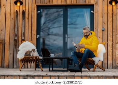 Mature man sitting on terrace reading book, enjoying cup of coffee during cold winter day. Peaceful moment alone in cabin. - Powered by Shutterstock