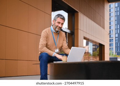 Mature man smiling at camera while working on laptop outdoors with a coffee