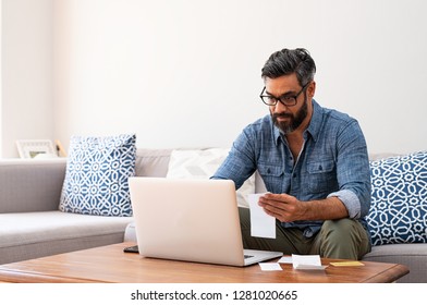 Mature man with spectacles and working on bills on laptop at home. Man with holding bills paying taxes with internet banking. Indian guy working on computer with invoice on table with copy space.