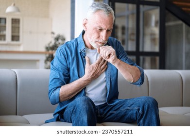 Mature man suffering from dry cough and chest pain while sitting alone on sofa in living room. Male feeling sick, has fever and respiratory infection symptoms. Healthcare concept - Powered by Shutterstock