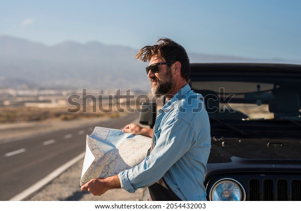Mature man in sunglasses searching direction on\
location map while standing beside bonnet of auto at highway.\
Handsome man reading map and looking away next to his car - male\
people travel lifestyle