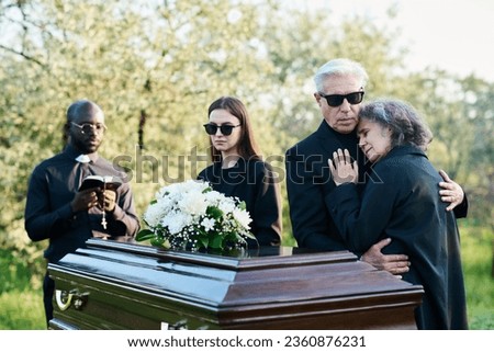 Mature man supporting his inconsolable wife while both grieving about their dead family member by coffin with closed lid at funeral service