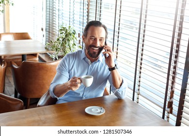 Mature man trader sitting at table at cafe at daytime holding cup drinking hot coffee talking with friend on smartphone laughing cheerful - Powered by Shutterstock