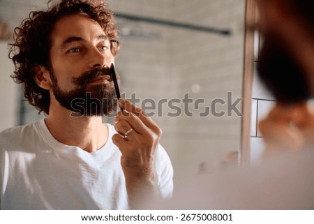 Mature man using a comb while styling his mustache in front of a mirror.