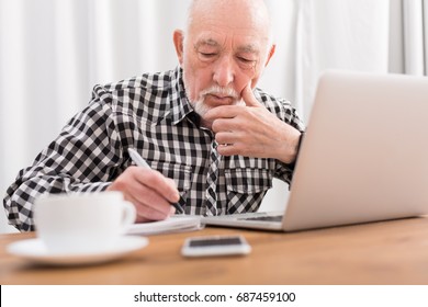 Mature man using laptop and writing in notepad at home desk. Education, recipe concept