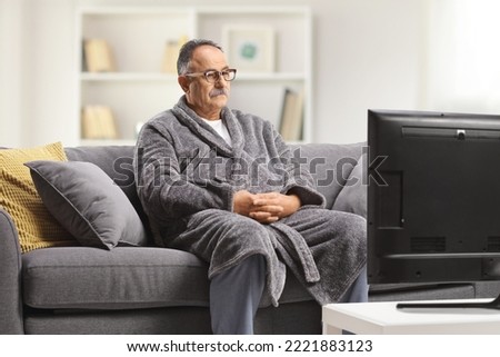 Mature man wearing a bathrobe, sitting on a gray sofa and watchin tv at home