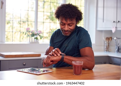 Mature Man Wearing Fitness Clothing At Home Logging Activity From Smart Watch Onto Digital Tablet