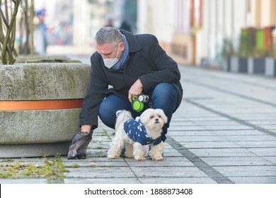 Mature man wearing a protective mask collects dog poop into a bag while walking down a city street